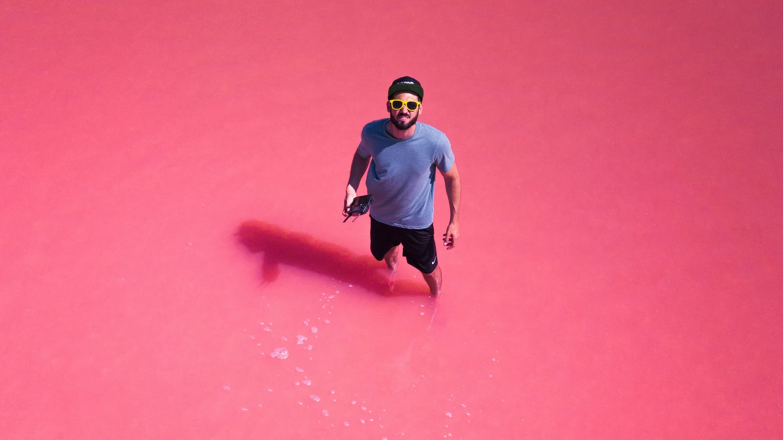 Homem de boné e óculos amarelos caminhando em água rosa, fotografado de cima.