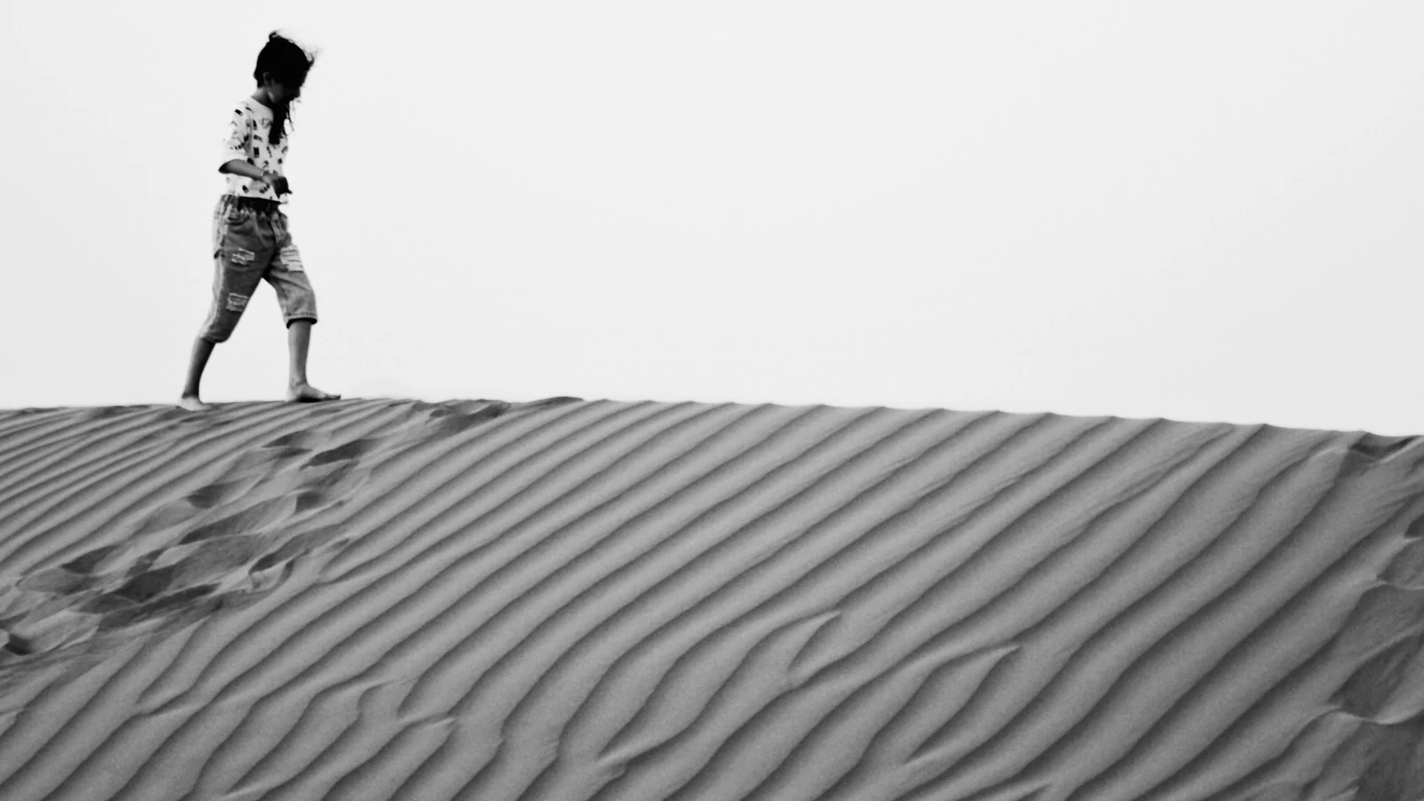 Pessoa caminhando descalça sobre uma duna de areia, em fotografia em preto e branco.