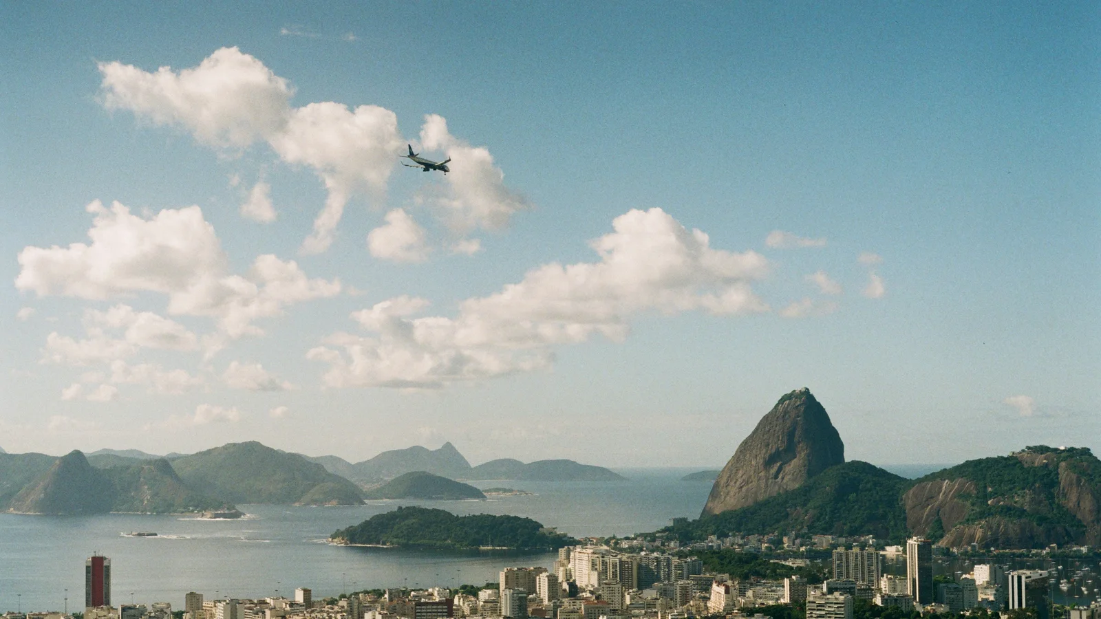 Vista do Rio de Janeiro com o Pão de Açúcar, a Baía de Guanabara e um avião no céu.