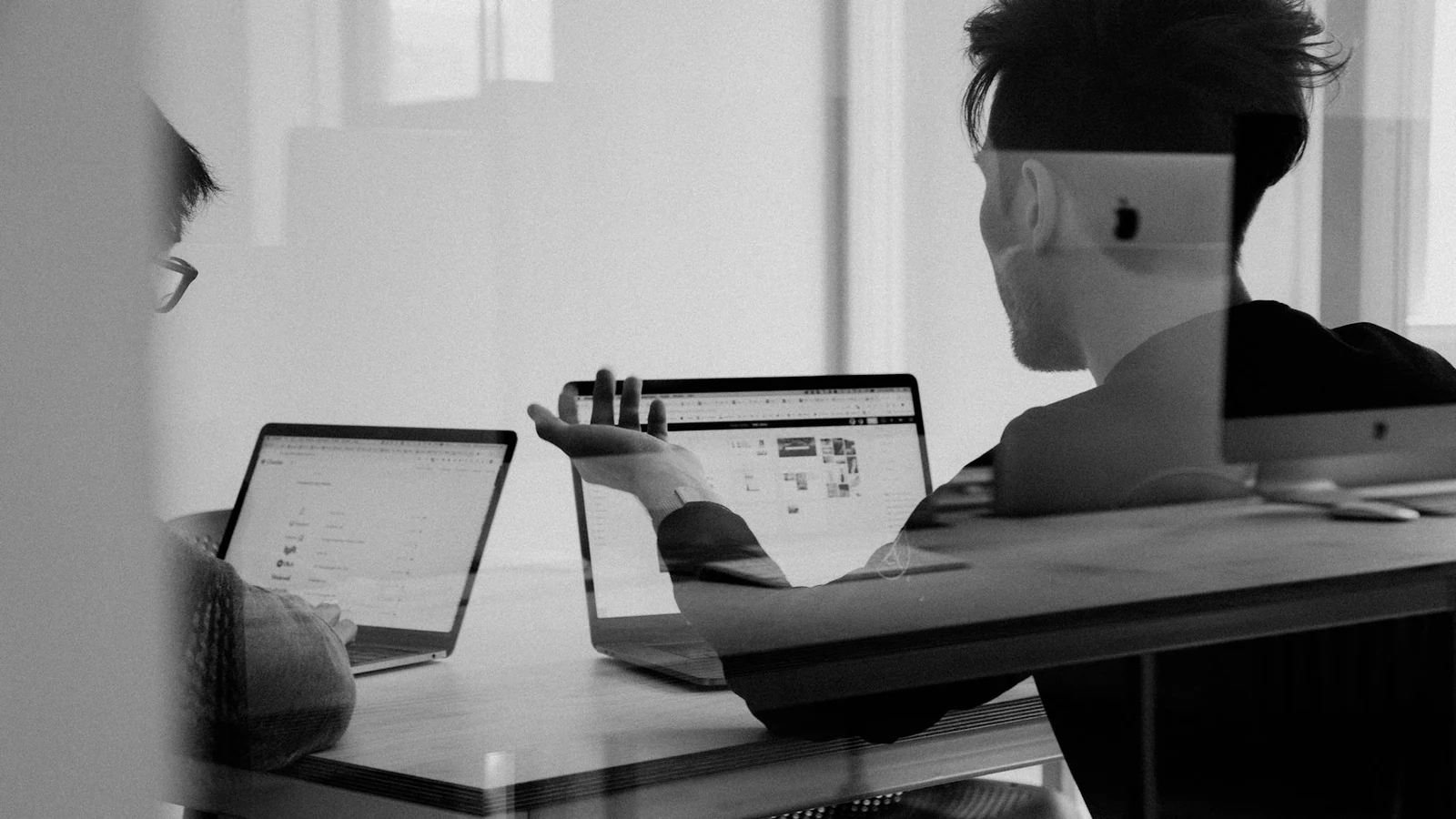 Duas pessoas conversando em uma mesa com notebooks, fotografadas em preto e branco através de uma divisória de vidro.