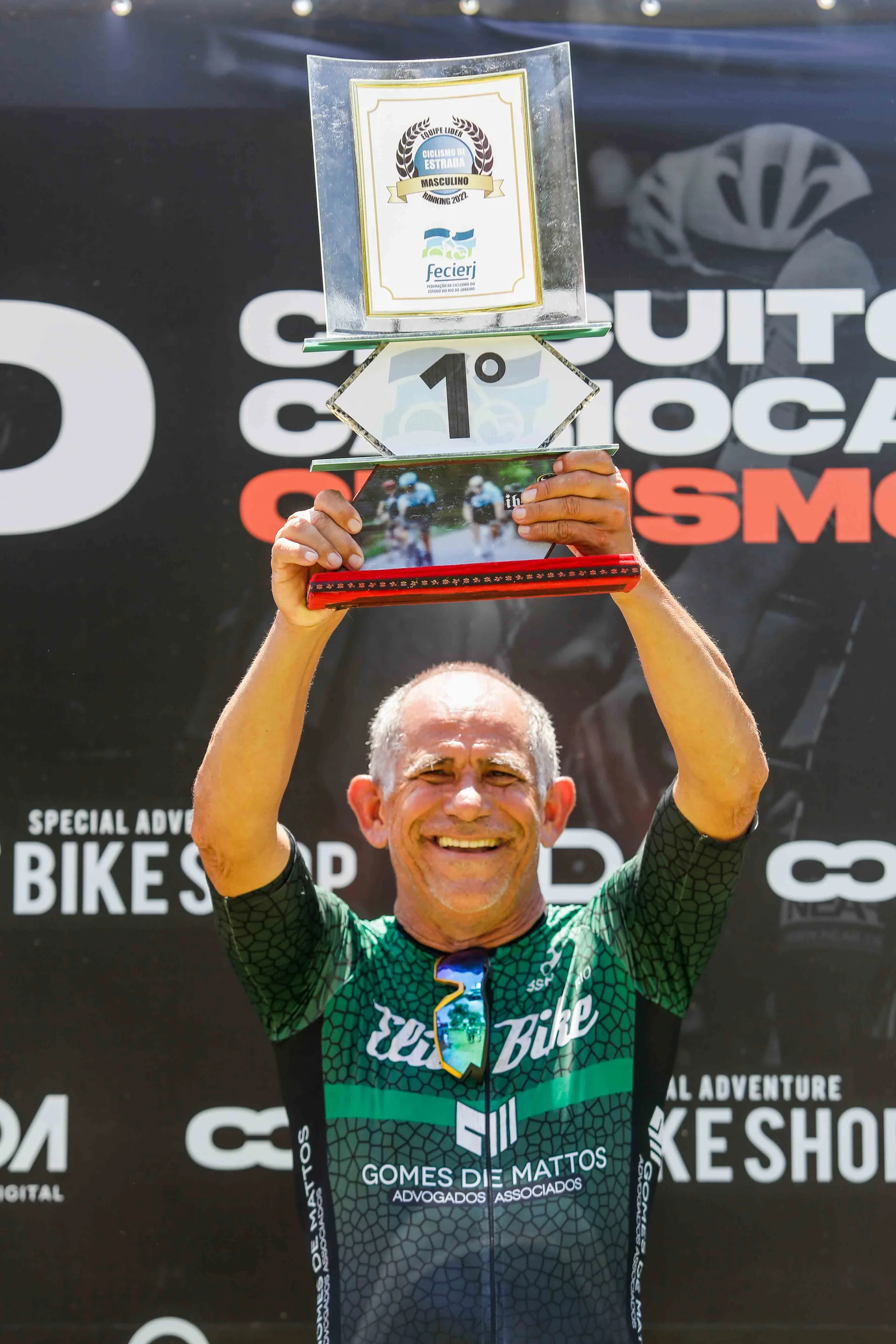 Ciclista ergue o troféu diante do painel do evento.
