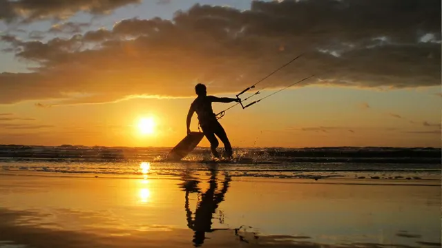 Praticante de kitesurf caminhando pela praia ao pôr do sol.