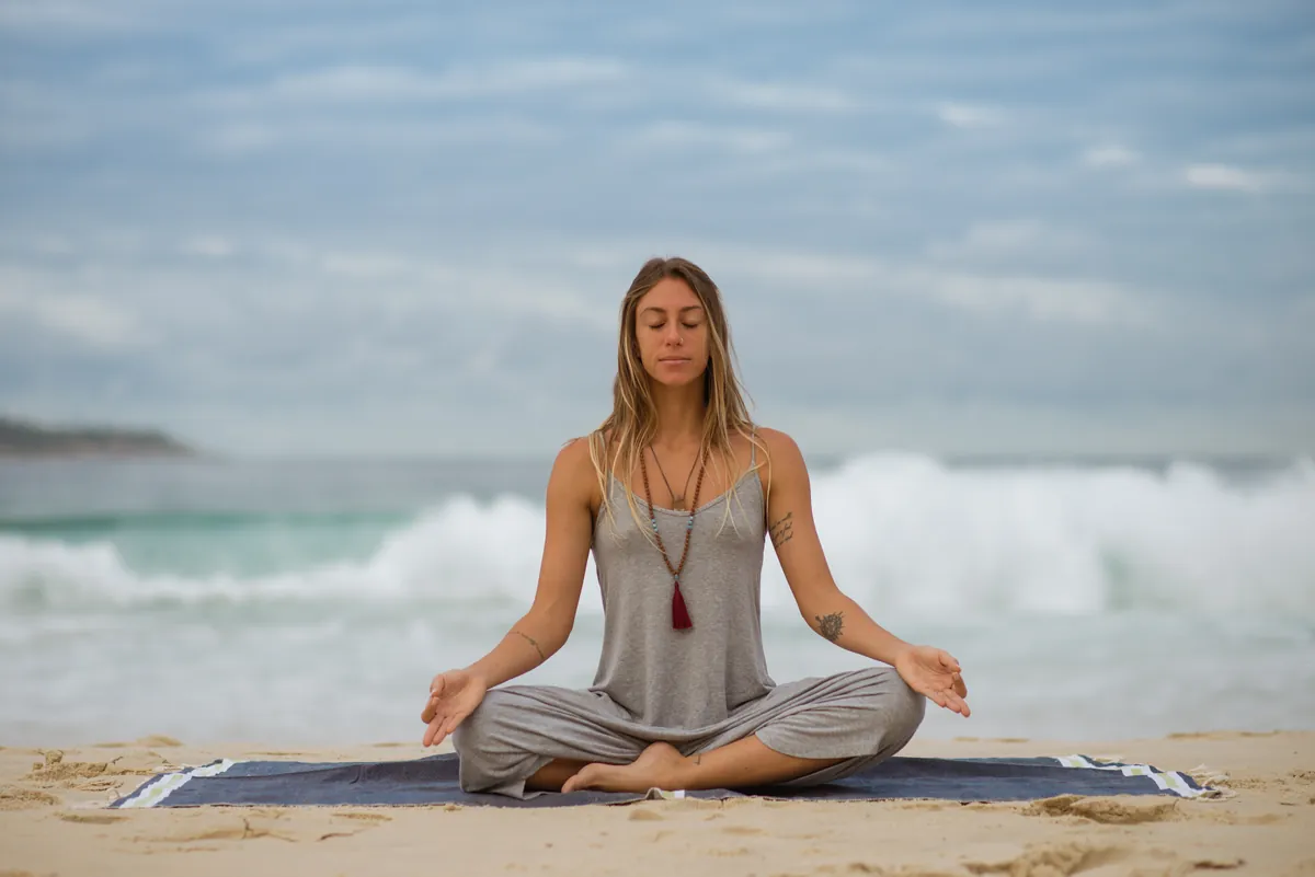 Mulher meditando sentada na areia, de frente para o mar.