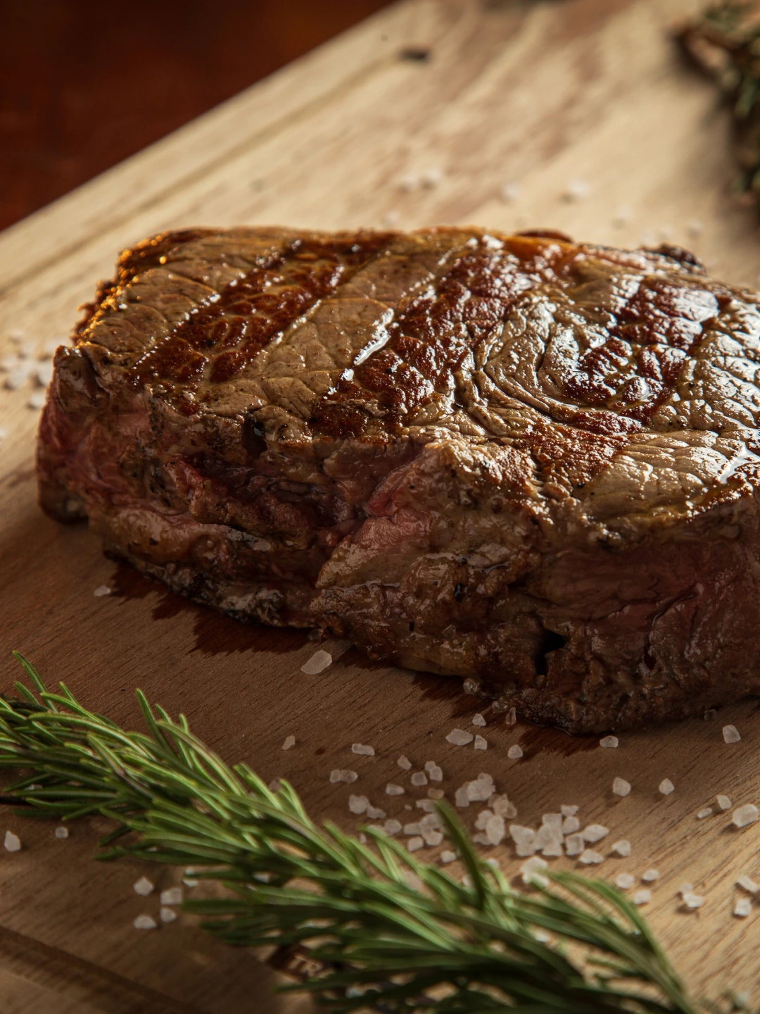 Carne grelhada servida sobre tábua de madeira com alecrim.