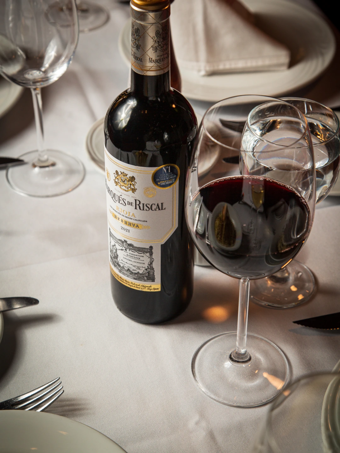 Garrafa de vinho tinto e taças em mesa posta com toalha branca.
