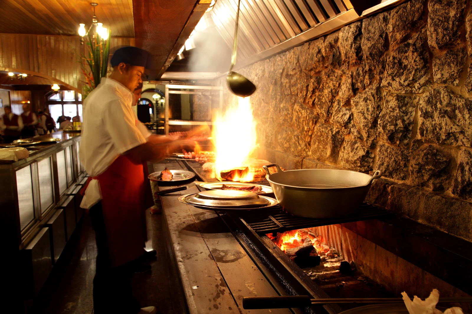 Cozinheiro junto à churrasqueira, com fogo, panelas e paredes de pedra.